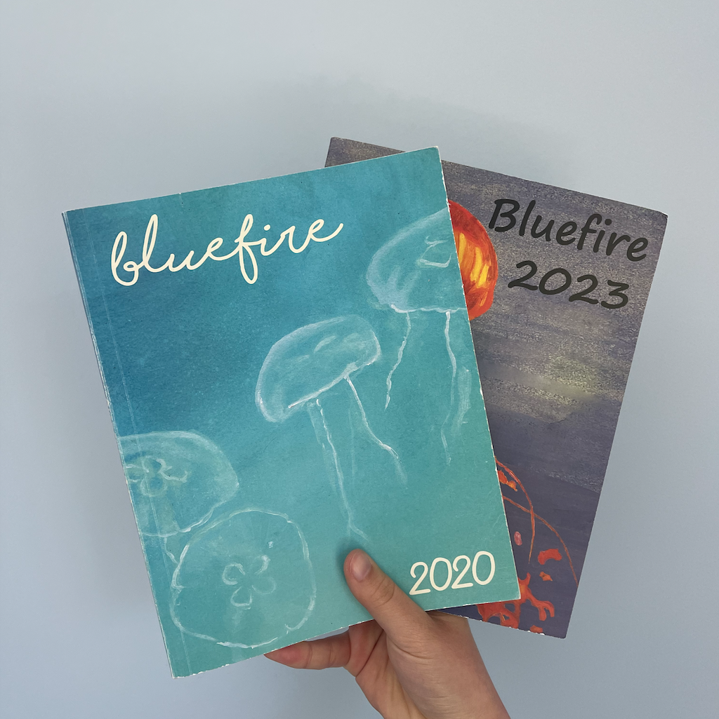 The Bluefire journal 2020 anthology and 2023 anthology held up in one hand against a blue background